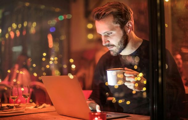 A guy at a restaurant at night sipping on a cup of coffee while on his computer.