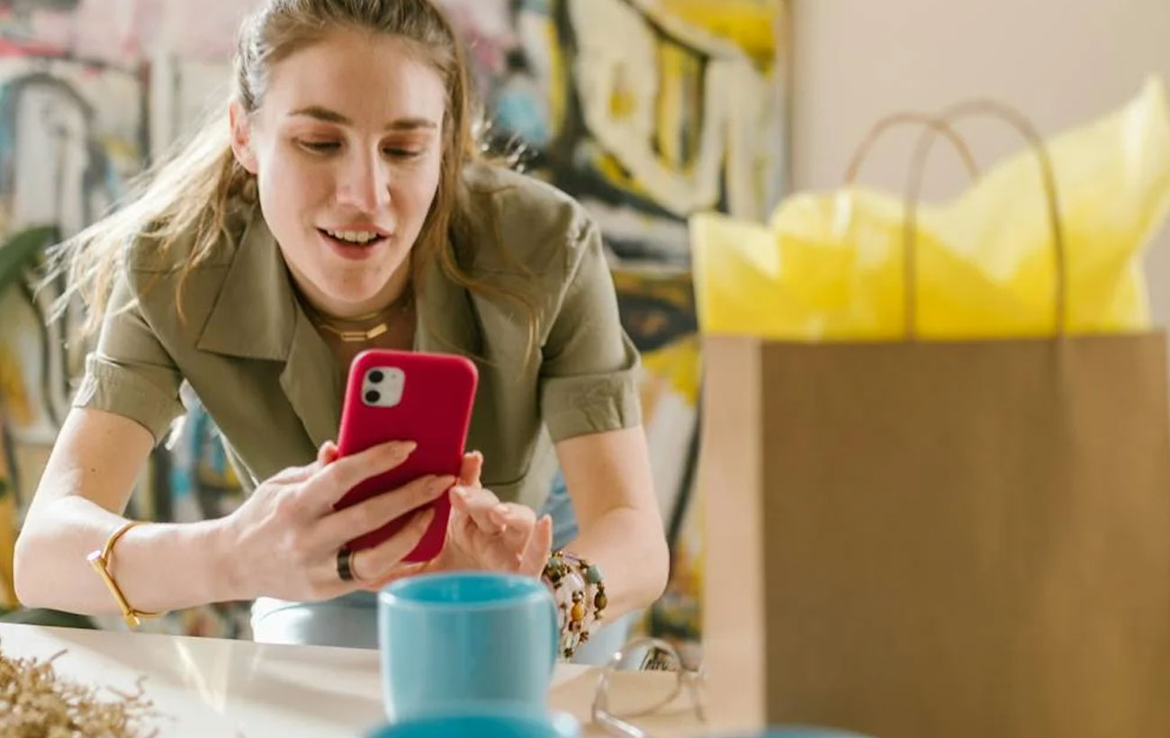 A lady at a coffee shop browsing on her red iPhone with a shopping bag in front of her.