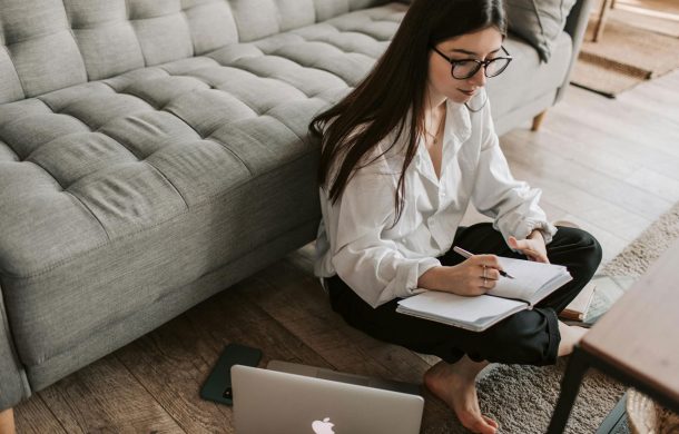 A lady sitting on the ground at home marking things down on her planner with her MacBook Pro and iPhone next to her.