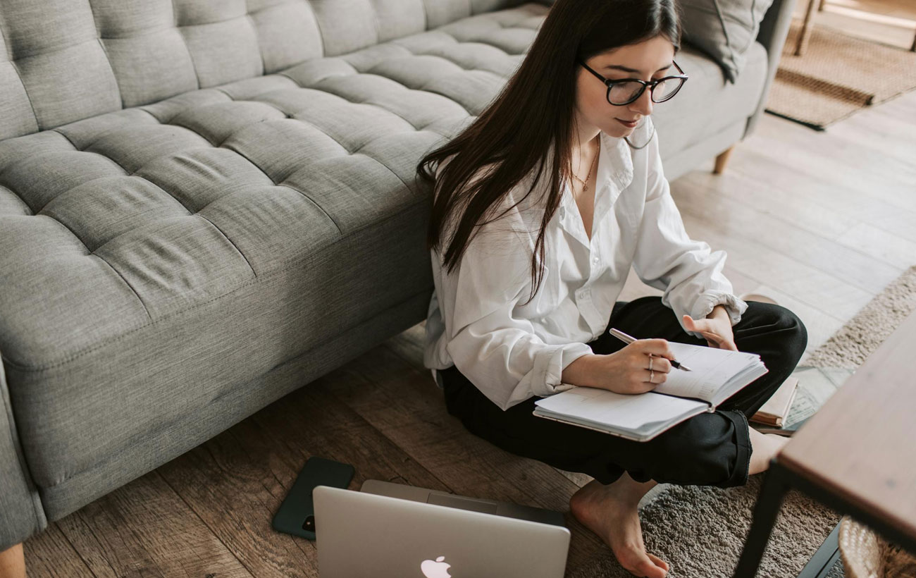 A lady sitting on the ground at home marking things down on her planner with her MacBook Pro and iPhone next to her.