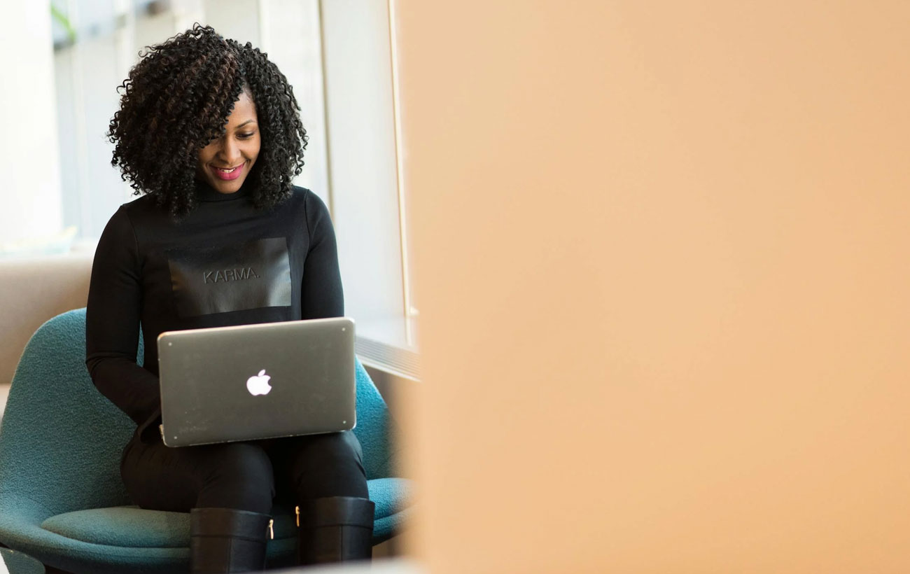 A lady with curly hair in all black sitting on a green chair browsing on her Macbook Pro.