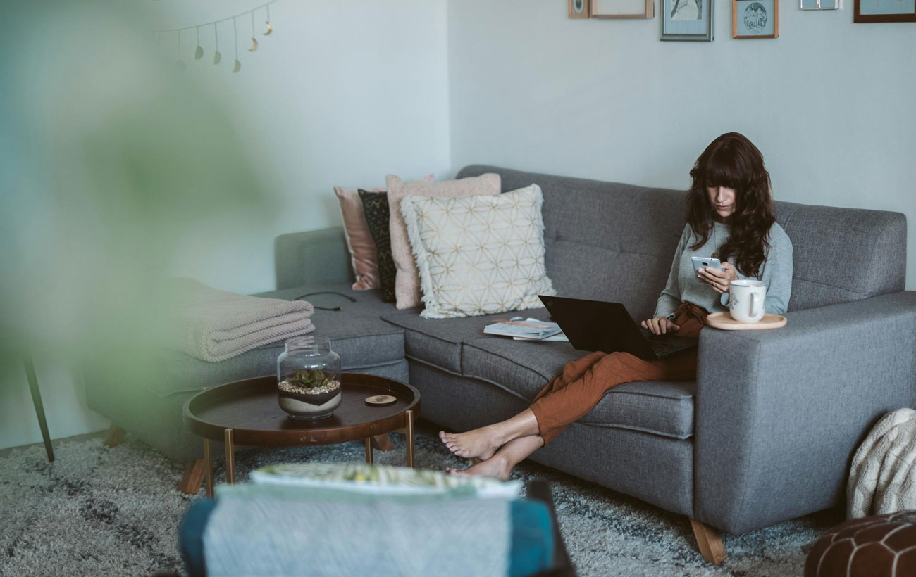 A brunette lady at home on the couch browsing her computer and phone.